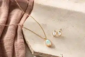 Goldene Kette mit Opalanhänger und zwei Opalohrstecker auf hellem Stein neben roséfarbenem Leinen