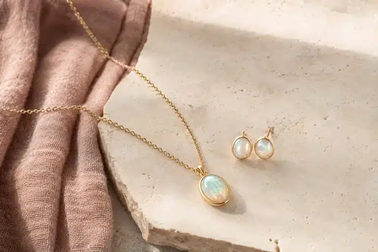 Goldene Kette mit Opalanhänger und zwei Opalohrstecker auf hellem Stein neben roséfarbenem Leinen